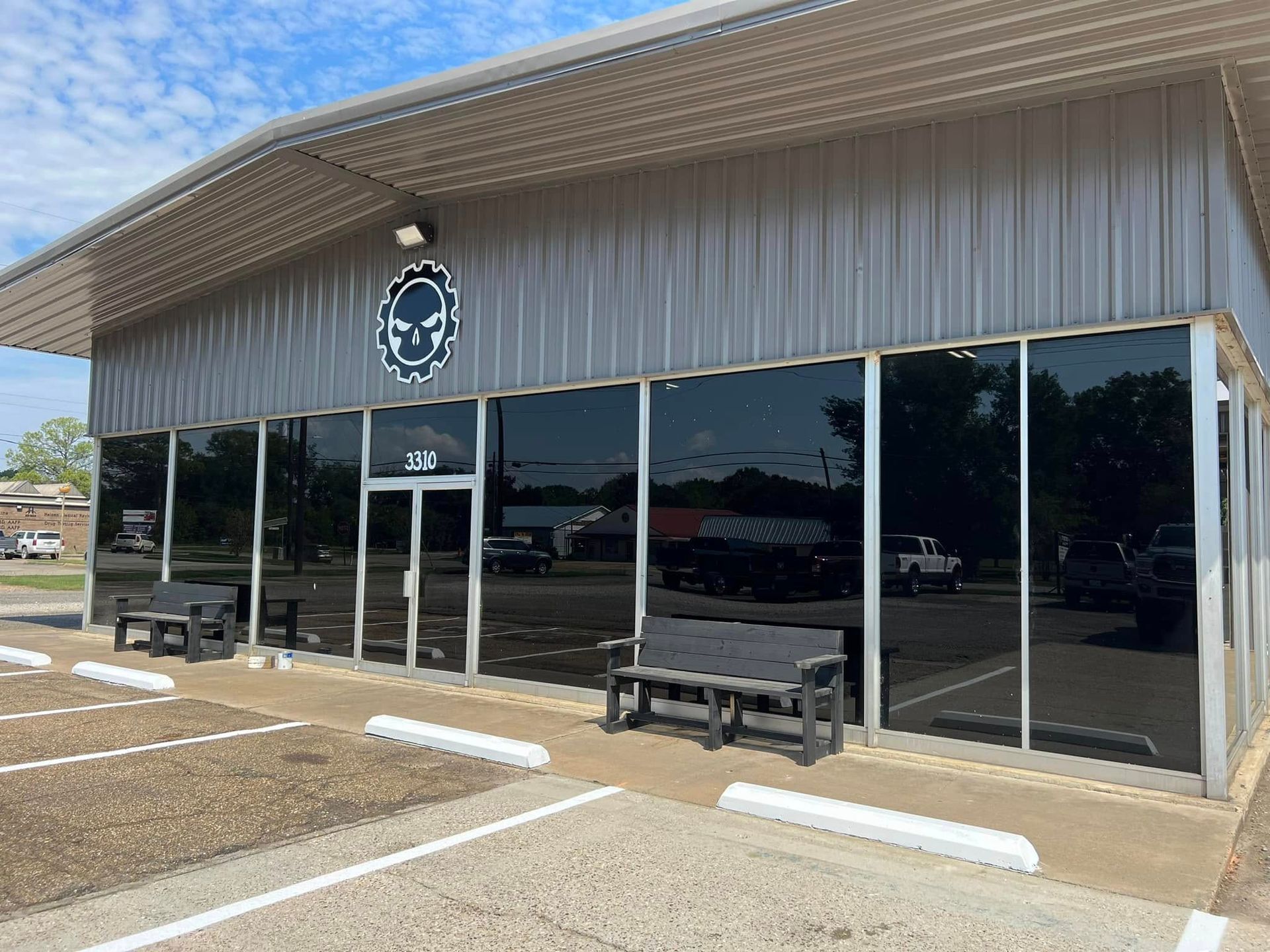 Storefront with large dark windows, a glass door, and benches in front of a metal-sided building.