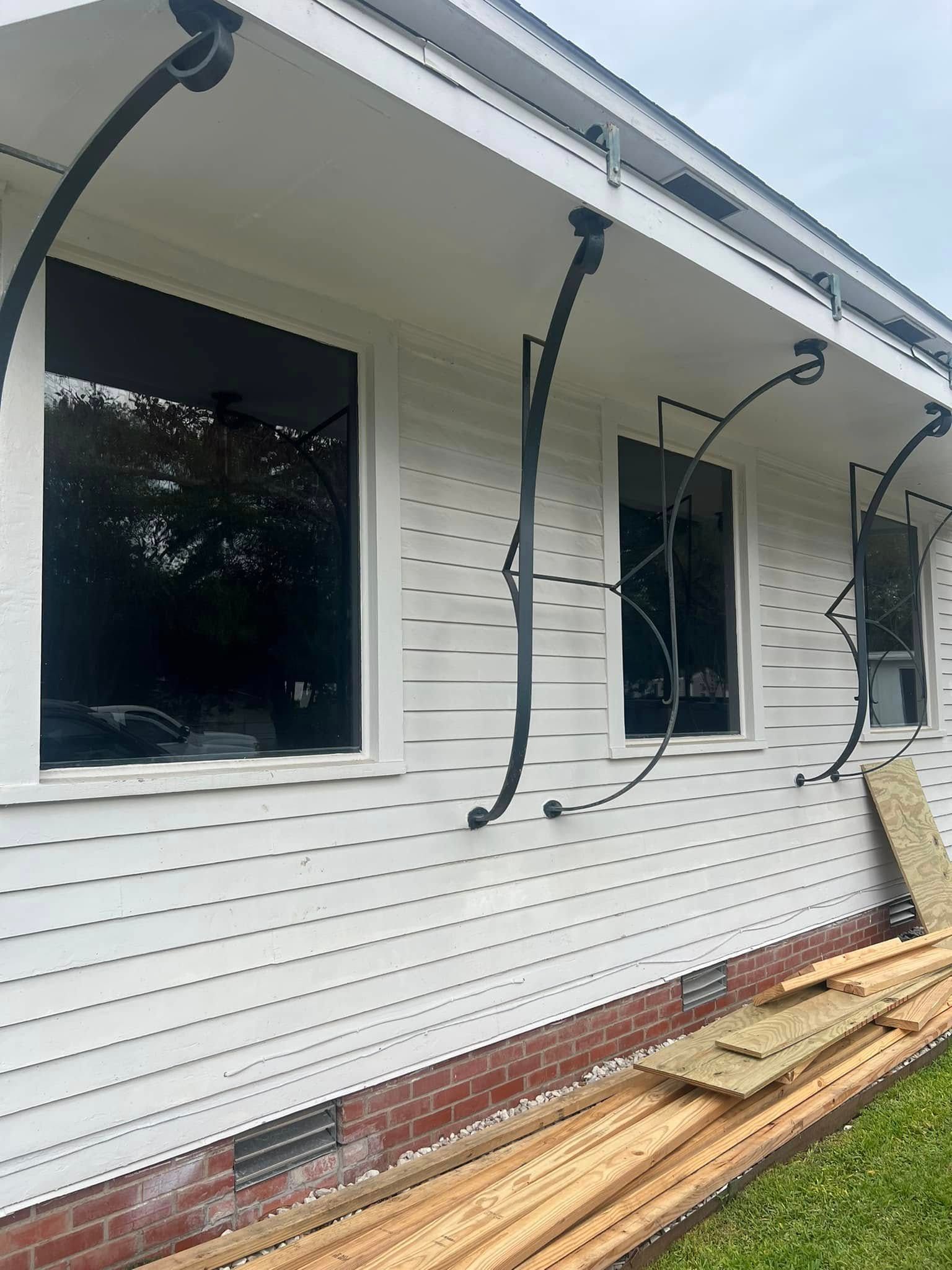 White house exterior with dark windows, decorative black awning brackets, and lumber stacked along the wall.