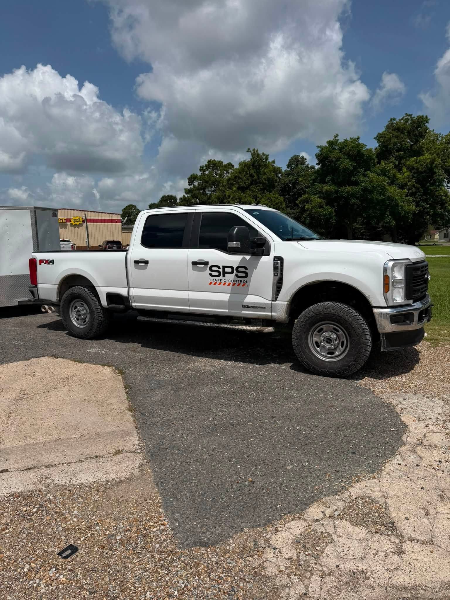 White utility pickup truck parked on gravel under cloudy skies, with trees and trailer in the background