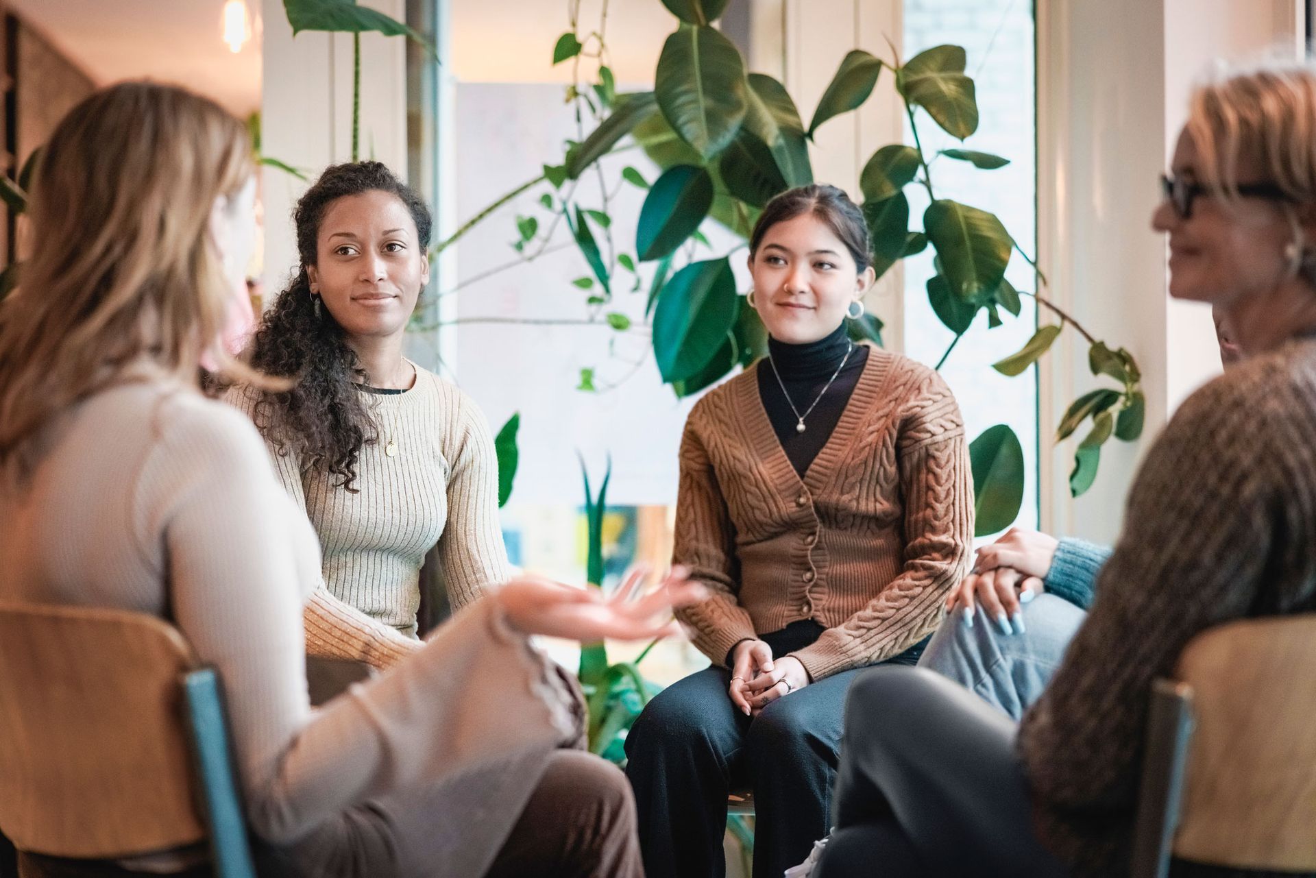Four people seated in a circle, engaged in conversation indoors. Plants visible.