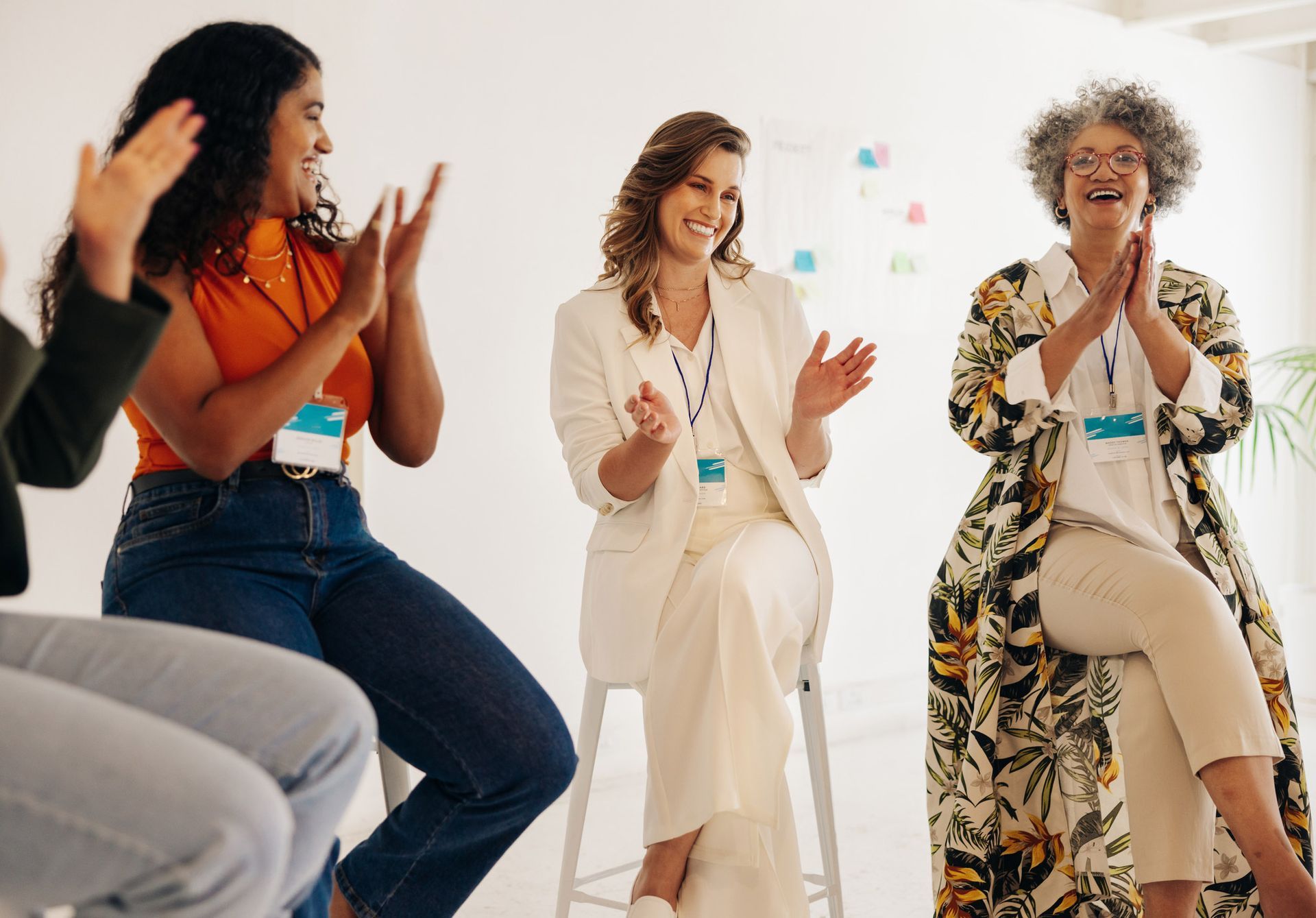 Three women seated in a circle, clapping and smiling. Light-filled room, white walls, name tags.