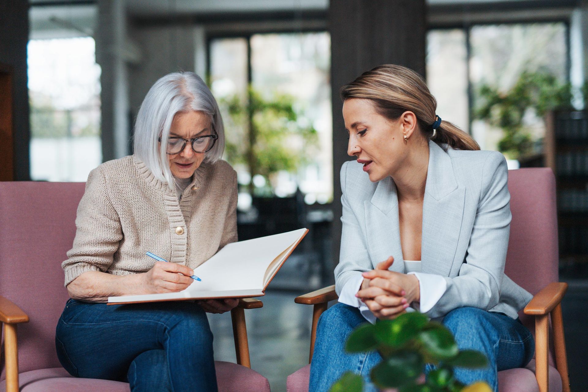 Two women in armchairs; one writing in notebook, the other looking on. Interior setting with plants.