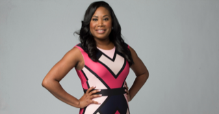 Woman in pink and black dress, hands on hips, smiling against a gray background.