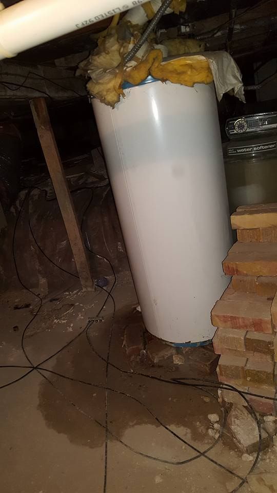 A leaking water heater sits on a concrete floor in a dimly lit crawl space next to a stack of bricks.