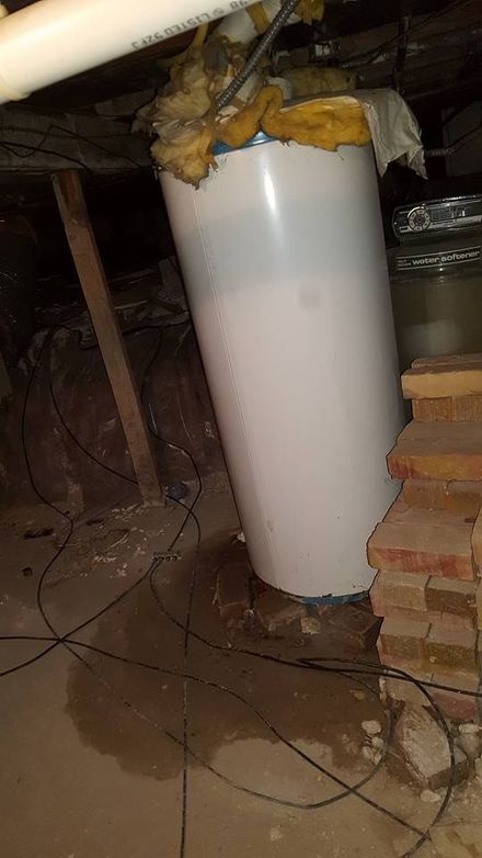 A leaking water heater sits on a concrete floor in a dimly lit crawl space next to a stack of bricks.