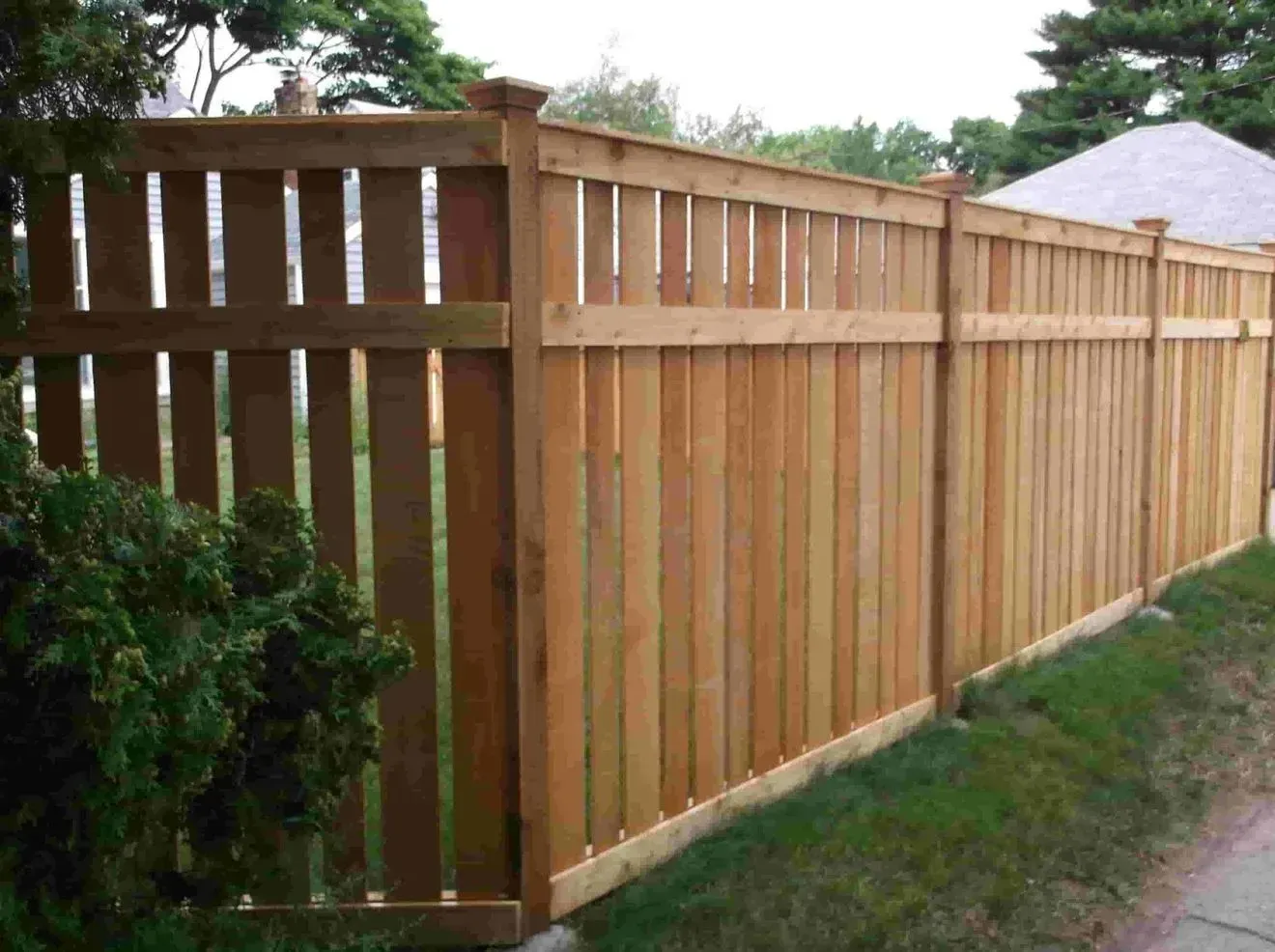 A wooden fence surrounds a lush green yard next to a sidewalk.