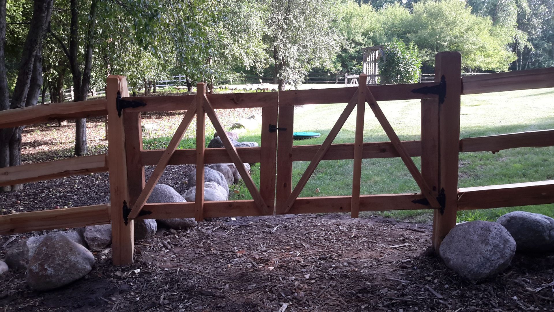 Wooden gate on a gravel path in a wooded park, with grass beyond and large rocks nearby.