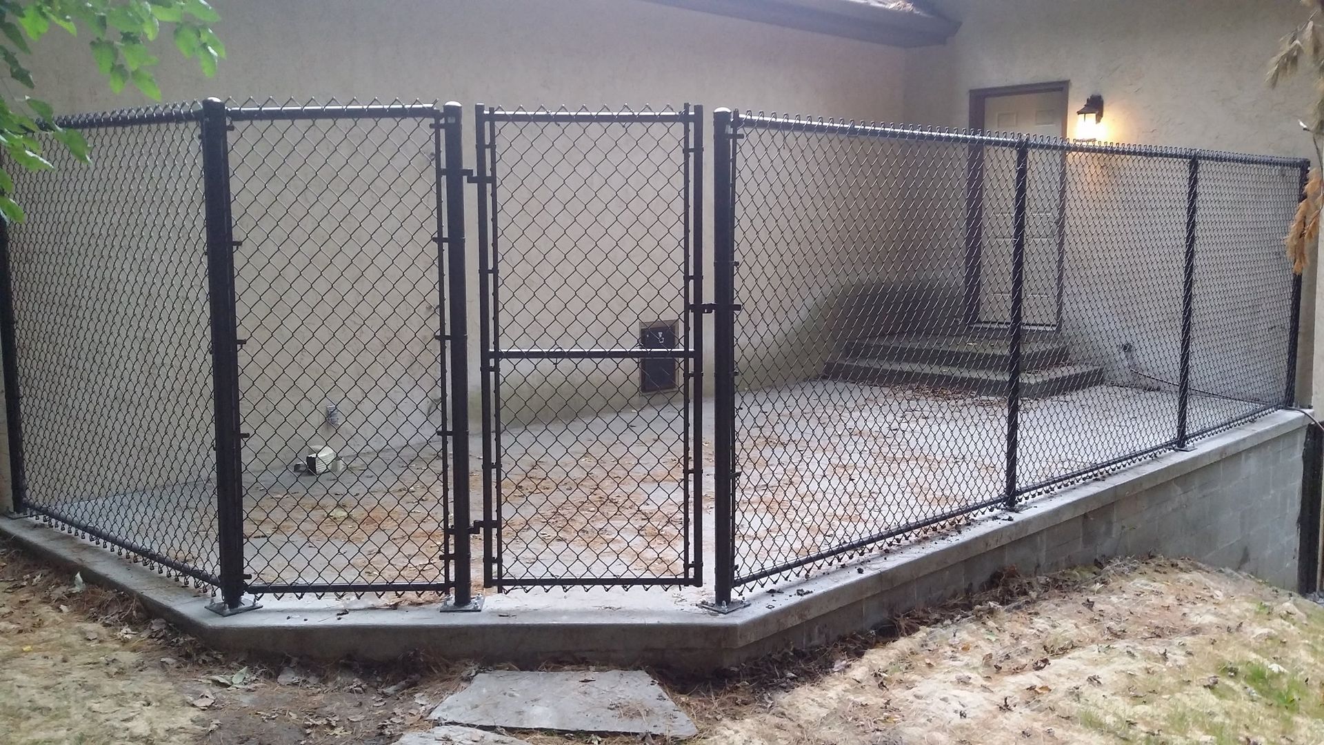 Chain-link kennel enclosure on a concrete pad beside a building, with a central gate and dirt ground nearby