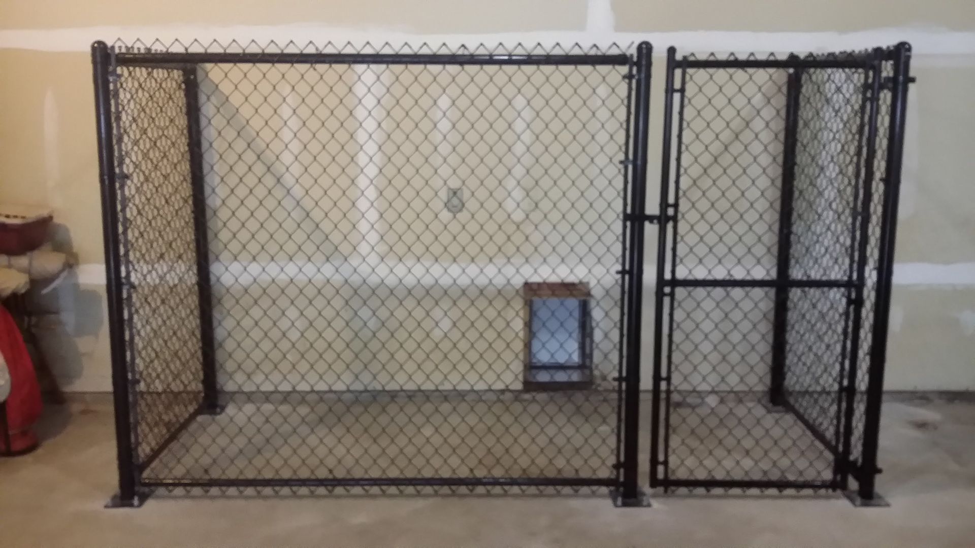 Black metal chain-link pet kennel with two sections in a beige room, with a small box inside.