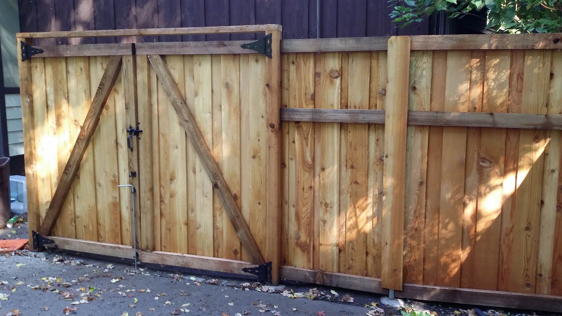 Wooden fence with a double gate and adjacent panel fence along a paved driveway, sunlit and slightly weathered