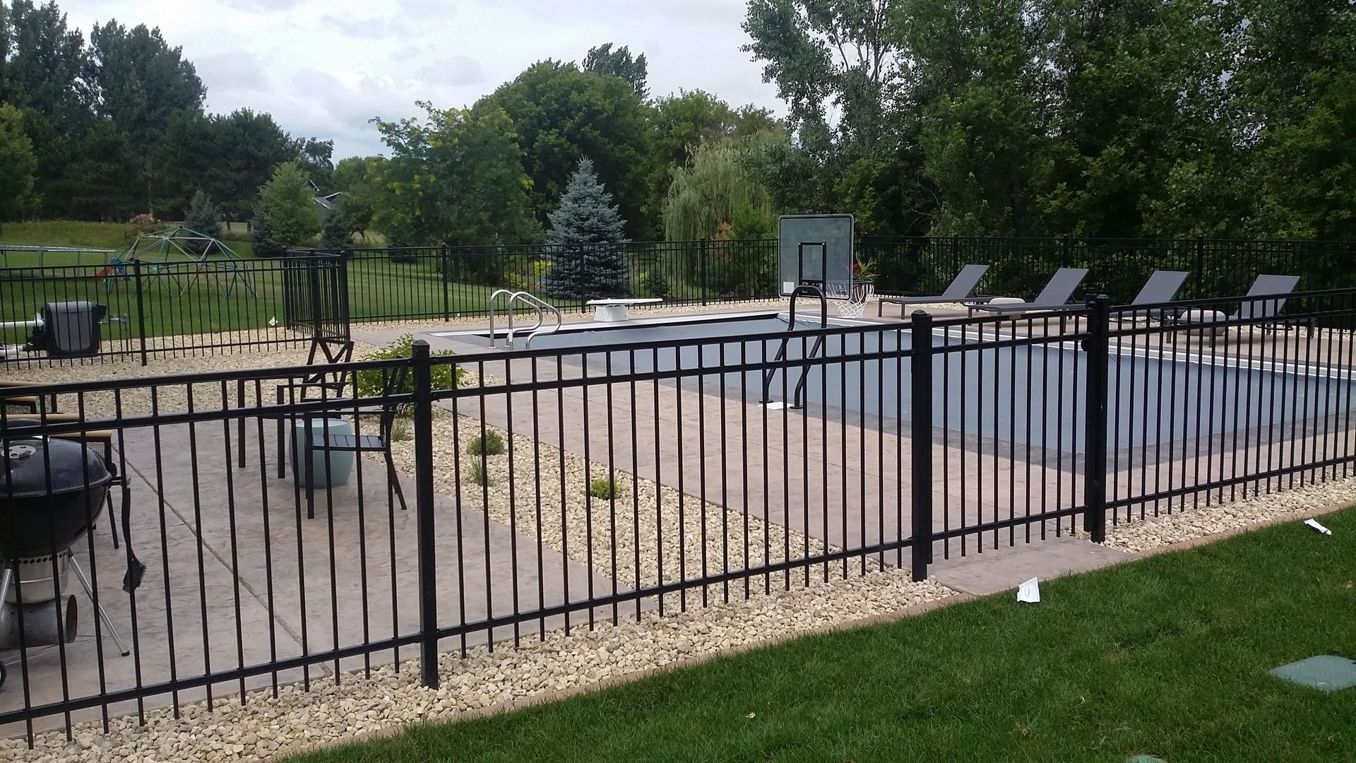 Black metal fence around an outdoor pool with lounge chairs, grassy lawn, trees, and a fountain in the background