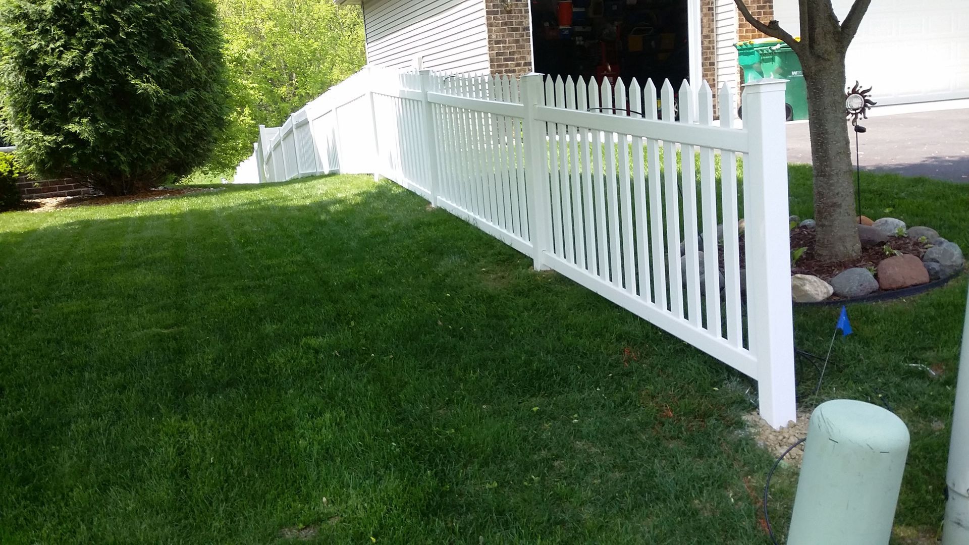 White picket fence along a grassy slope beside a house and garden.