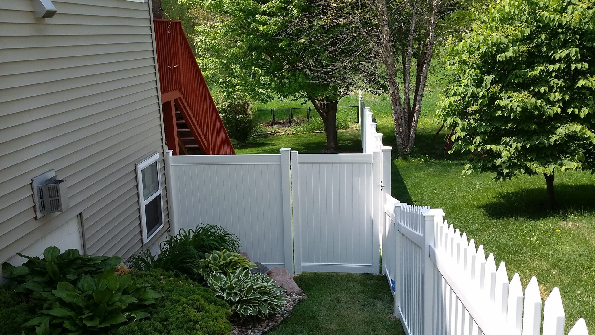 Backyard with white fence, house siding, red stairs, and green trees and lawn
