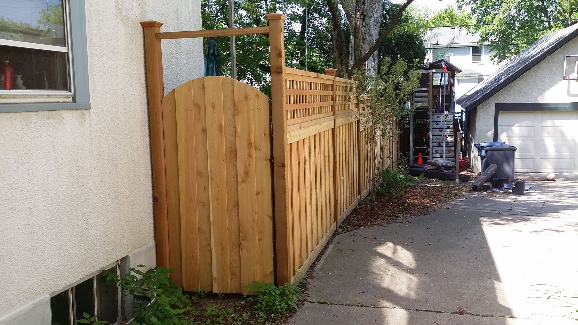 Wooden privacy fence beside a house along a concrete driveway in bright sunlight.