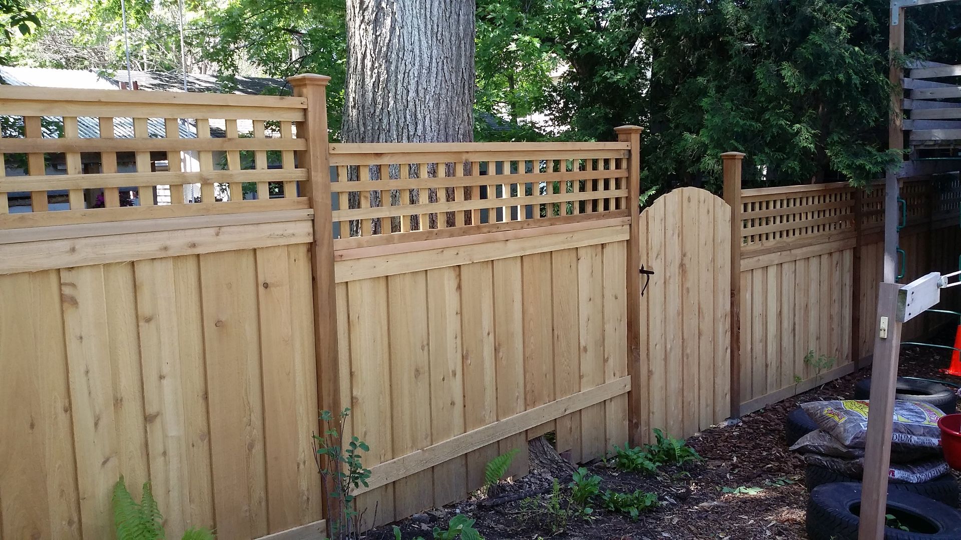 Wooden privacy fence with lattice top along a backyard, bordered by trees and garden beds.