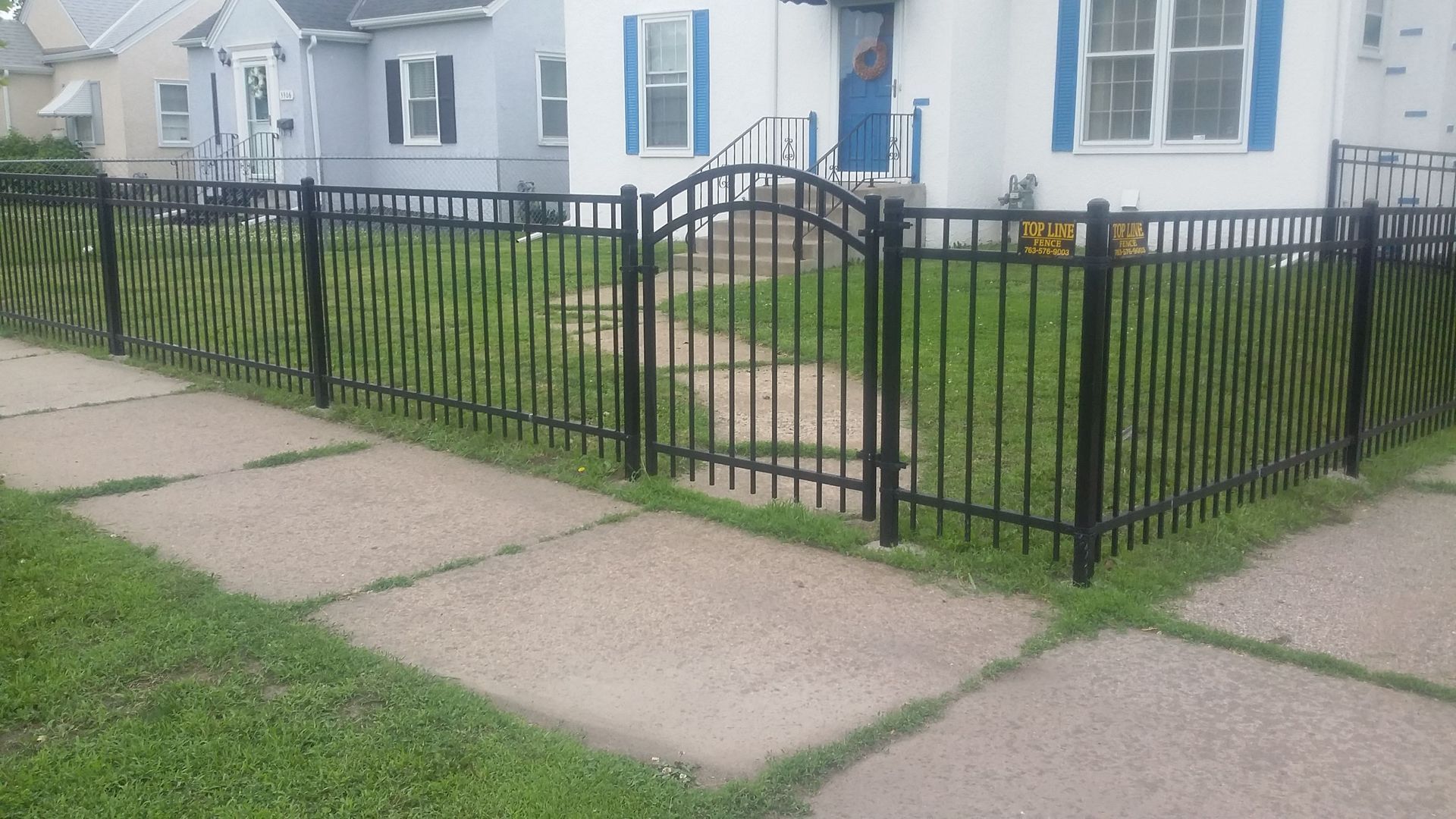 Black metal fence and arched gate around a white house with blue door and small front yard