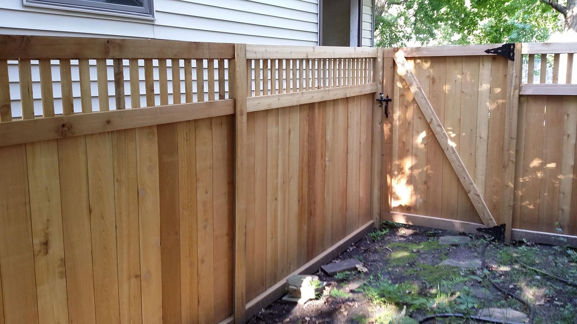Wooden backyard fence with gate beside a house, enclosing a small dirt and grass area.