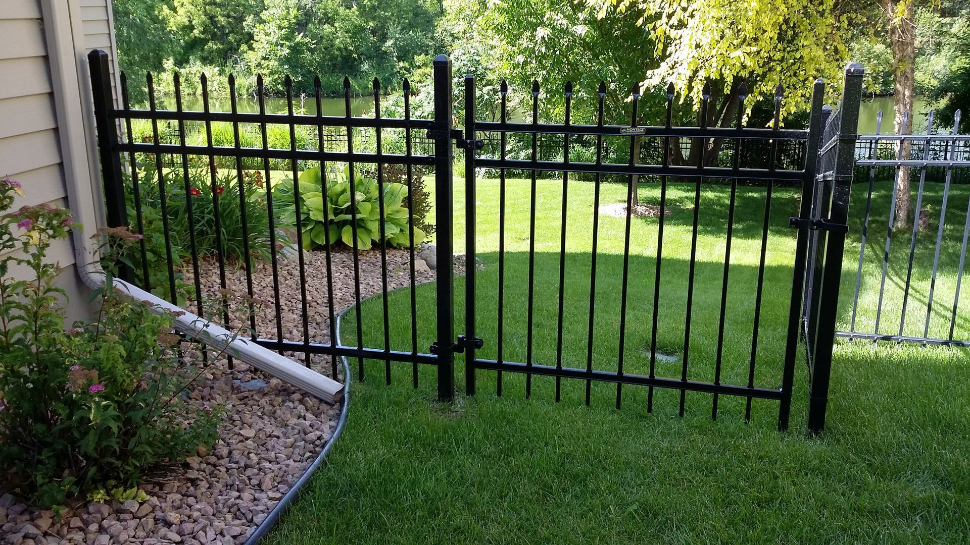 Black metal garden gate beside a beige house, opening onto a green lawn and trees.