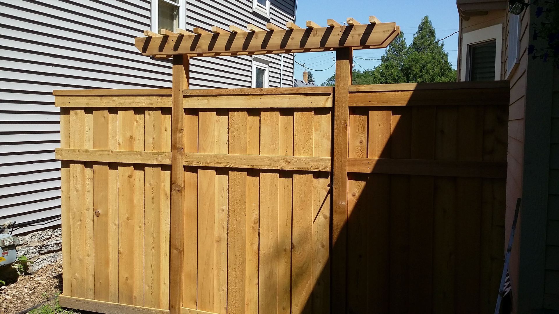 Wooden backyard fence gate with a striped pergola top beside a house and shed