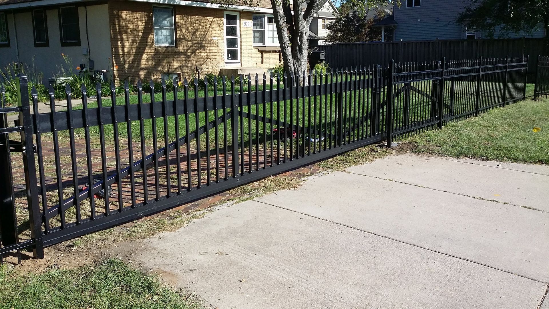 Black metal fence beside a concrete sidewalk in front of a house and lawn.