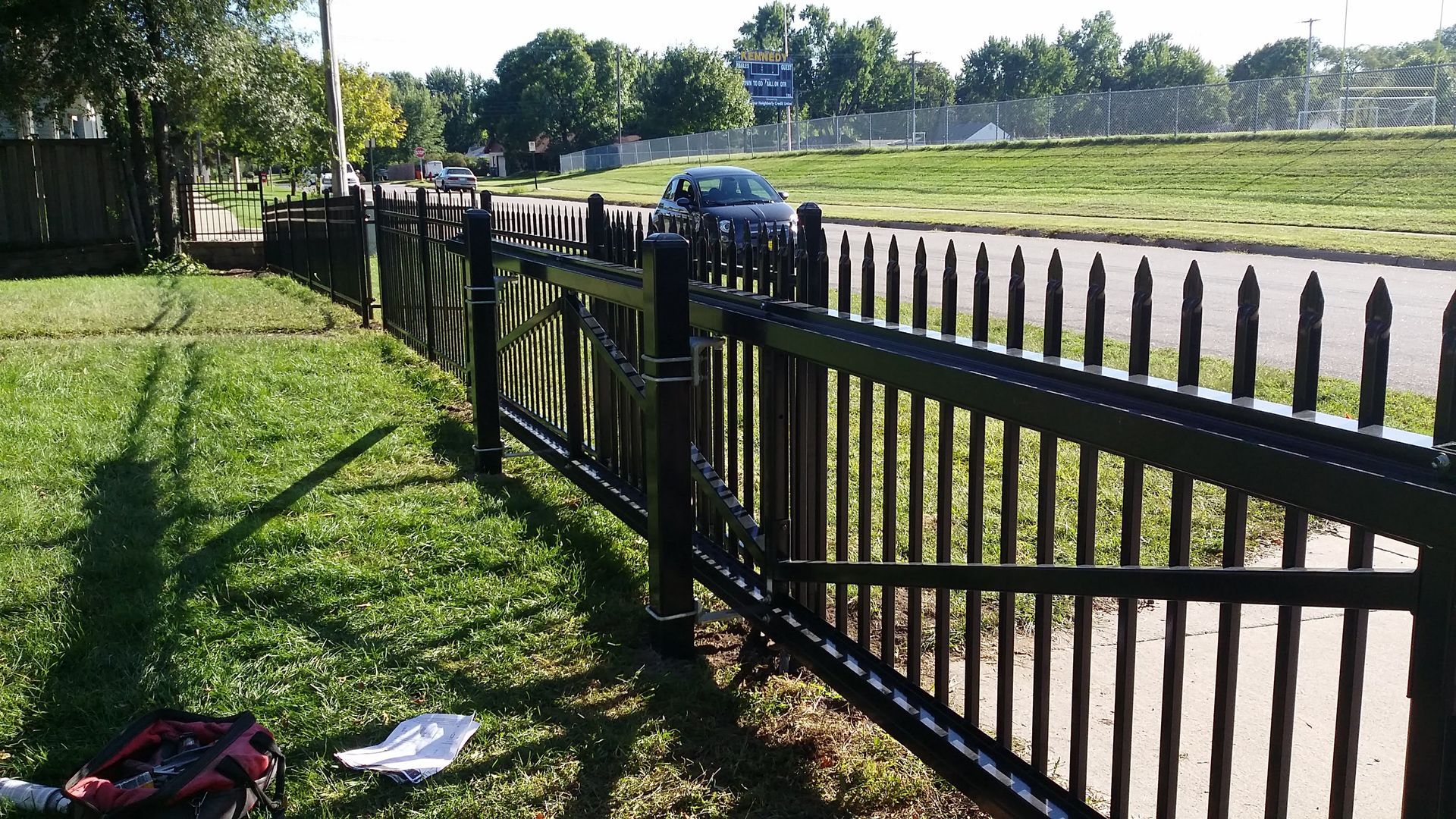 Black fence along a sunny roadside beside a grassy field, with a parked car in the distance