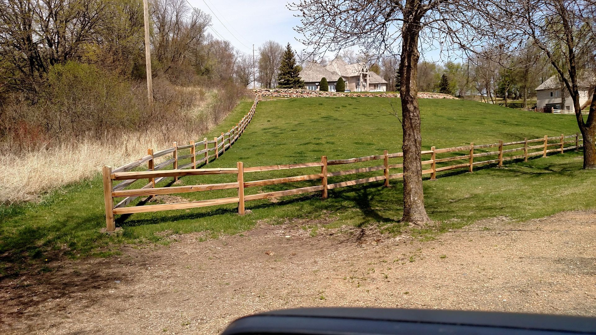 Grassy rural field with a wooden fence, trees, and a distant house on a cloudy day