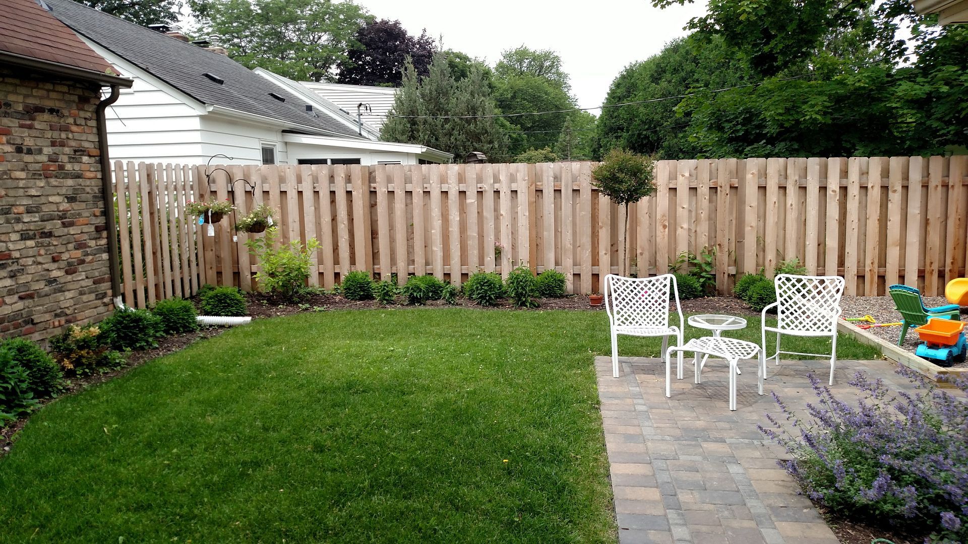 Backyard patio with green lawn, wooden fence, stone path, and white outdoor chairs around a small table