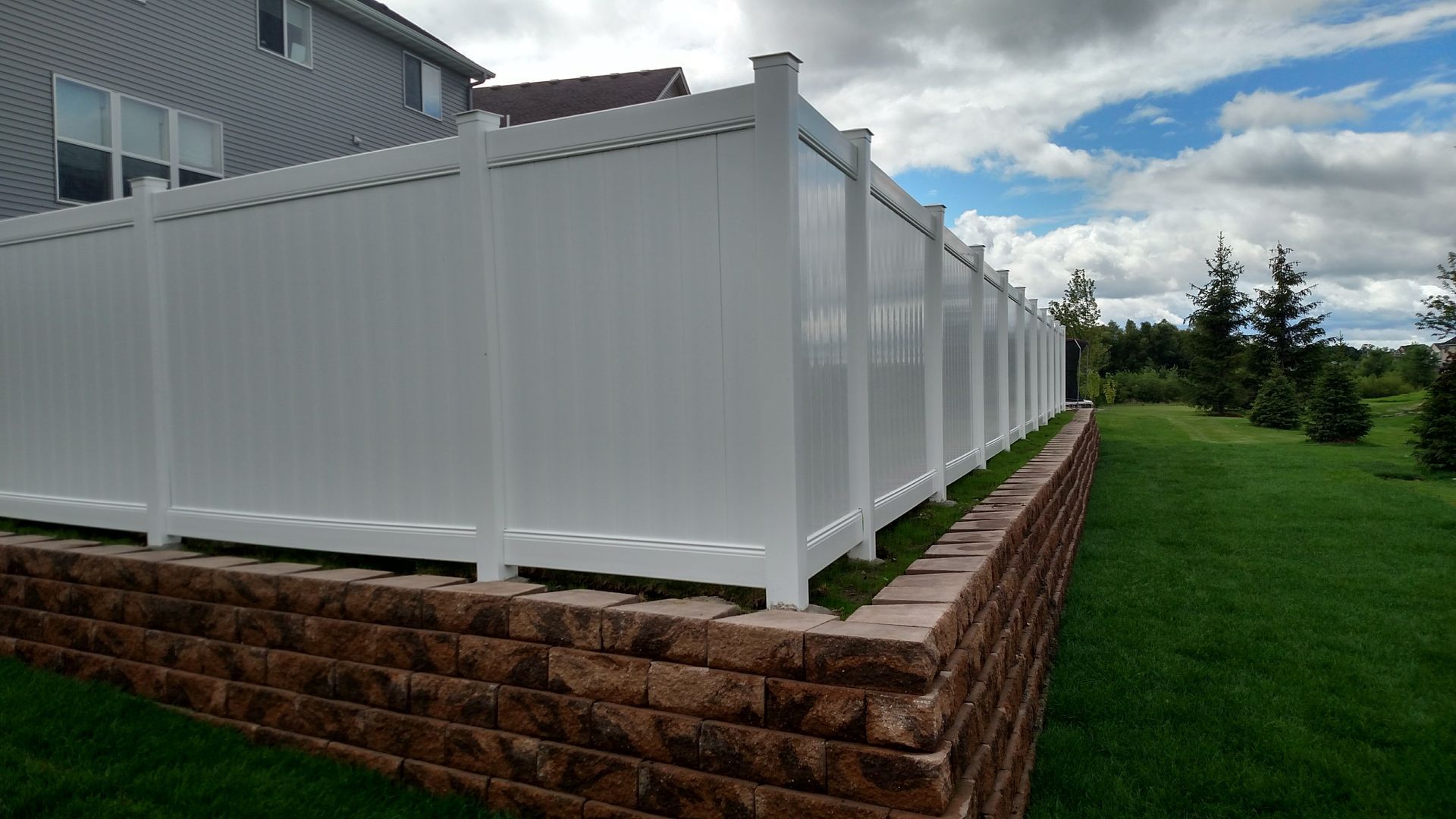 White vinyl fence along a brick retaining wall beside a green lawn and house under cloudy sky