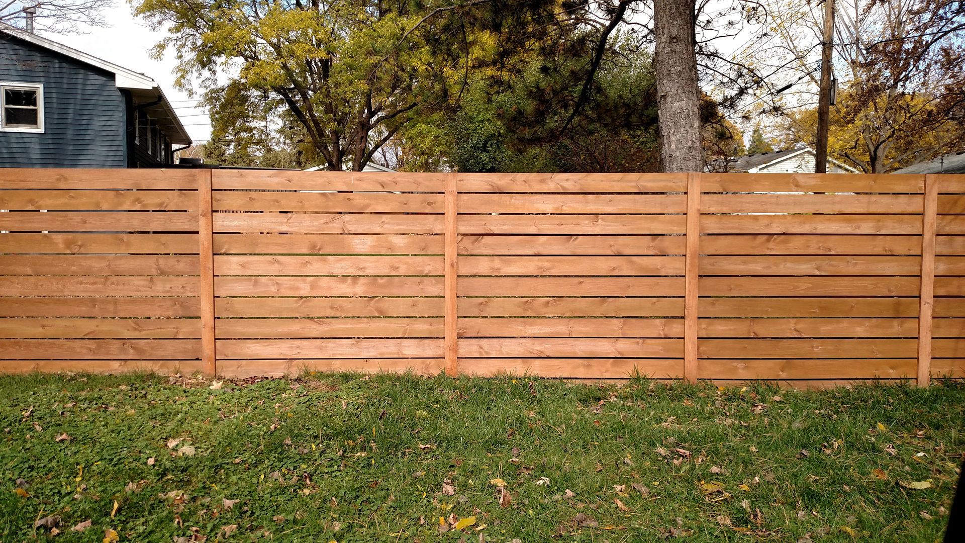 Wooden privacy fence across a grassy yard with autumn trees and a house in the background