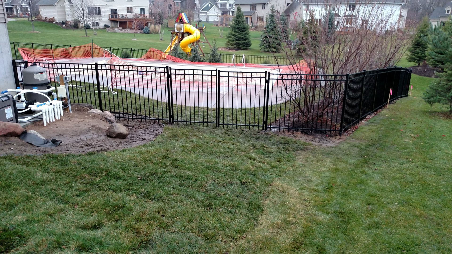 Backyard pool with black fence and a yellow slide, surrounded by grass and shrubs