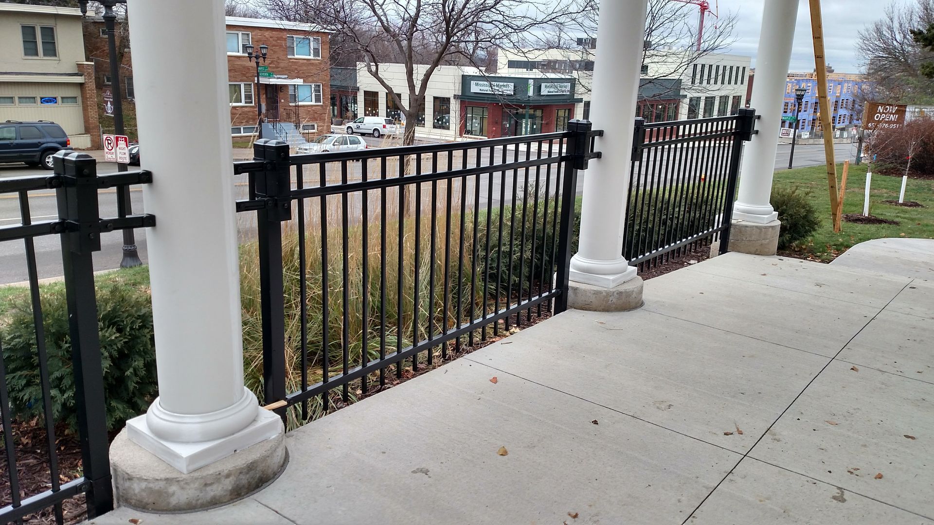 Black metal fence and white columns along a sidewalk by brick buildings in a neighborhood.