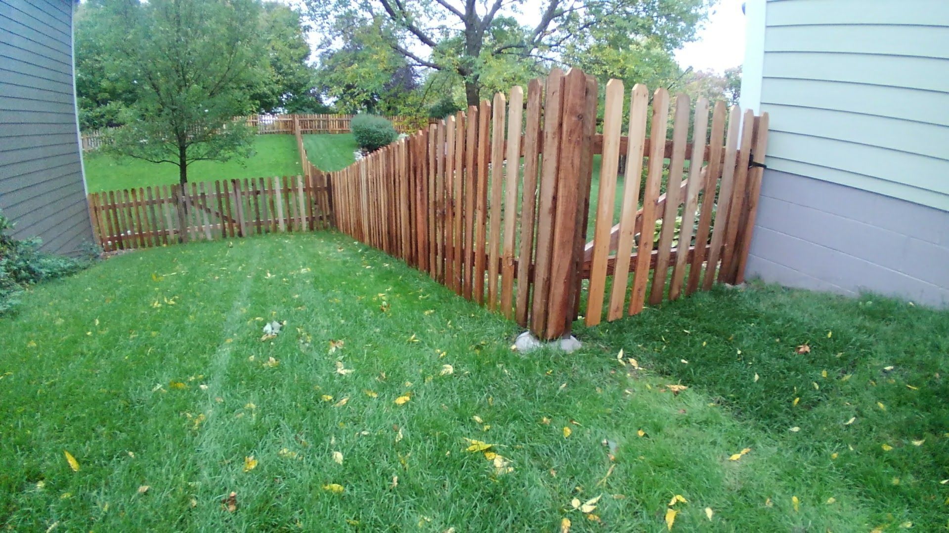 Wooden fence corner in a grassy backyard beside a house, with trees and a green fence in the background