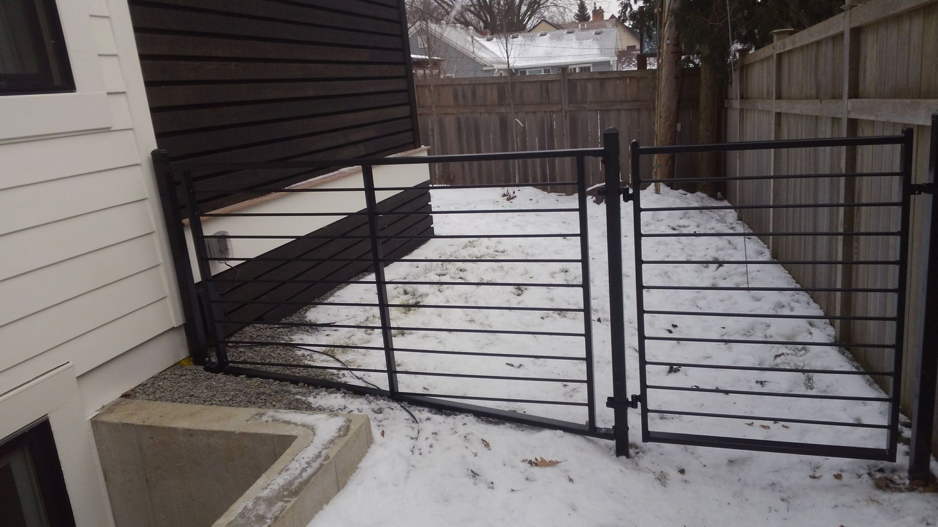 Snowy backyard patio with a black metal gate and fence beside a house