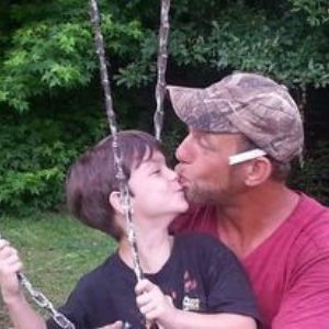 Two people sharing a kiss on a backyard swing, with green trees in the background.