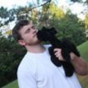 Man in a white shirt holding a black dog outdoors with trees in the background
