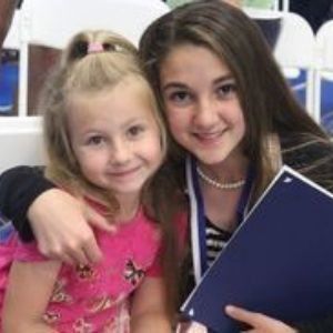 Two smiling girls pose together, one holding a blue folder, in a classroom setting.