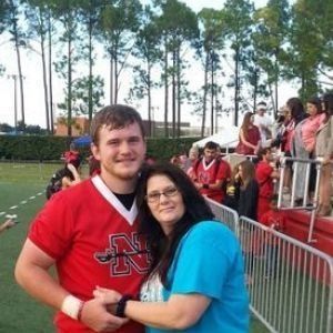 Two people in red and blue stand hugging on a football field near bleachers and trees.