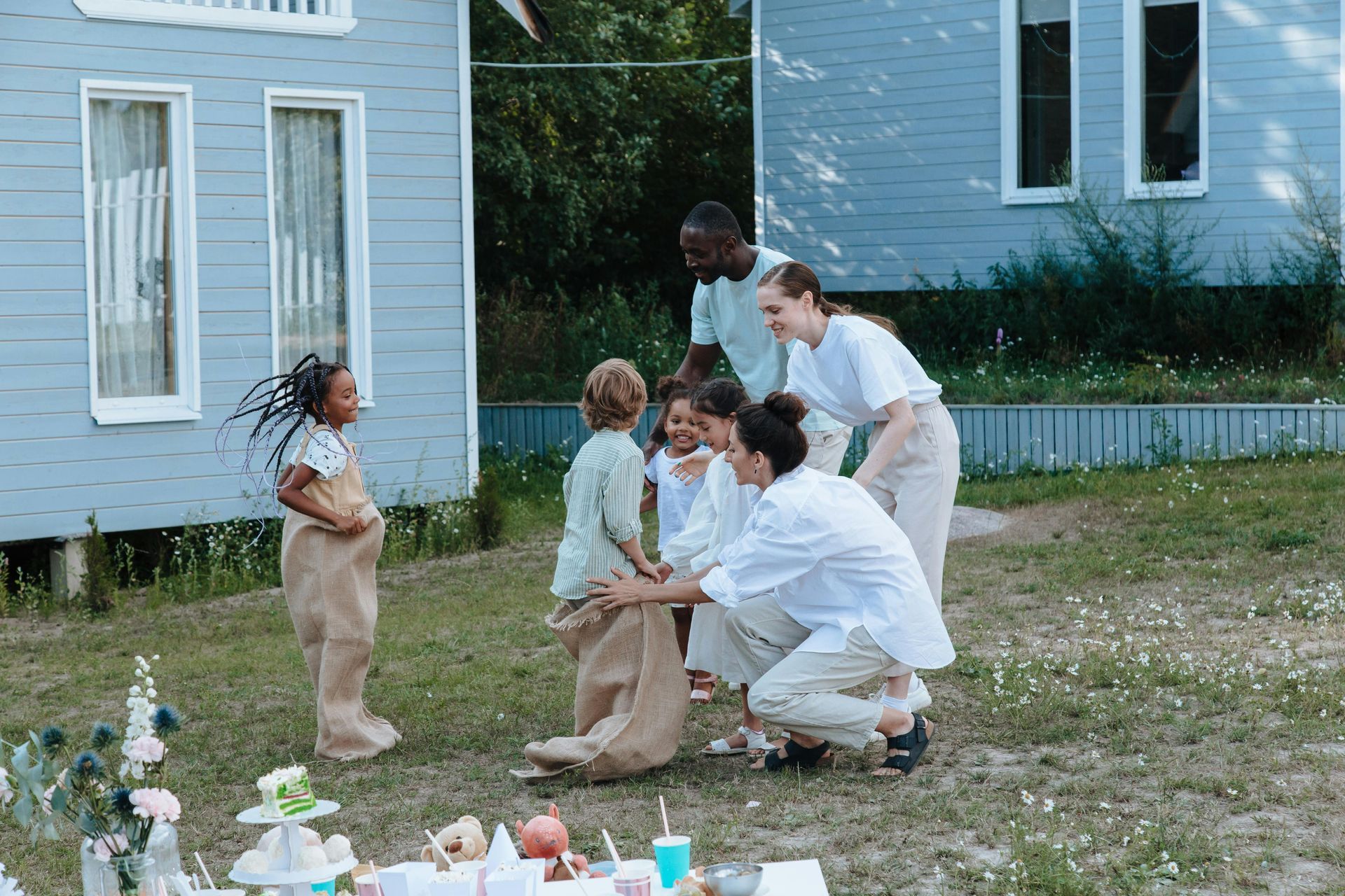 Children and adults gathered in a yard outside a blue house, crouching around small items on the ground