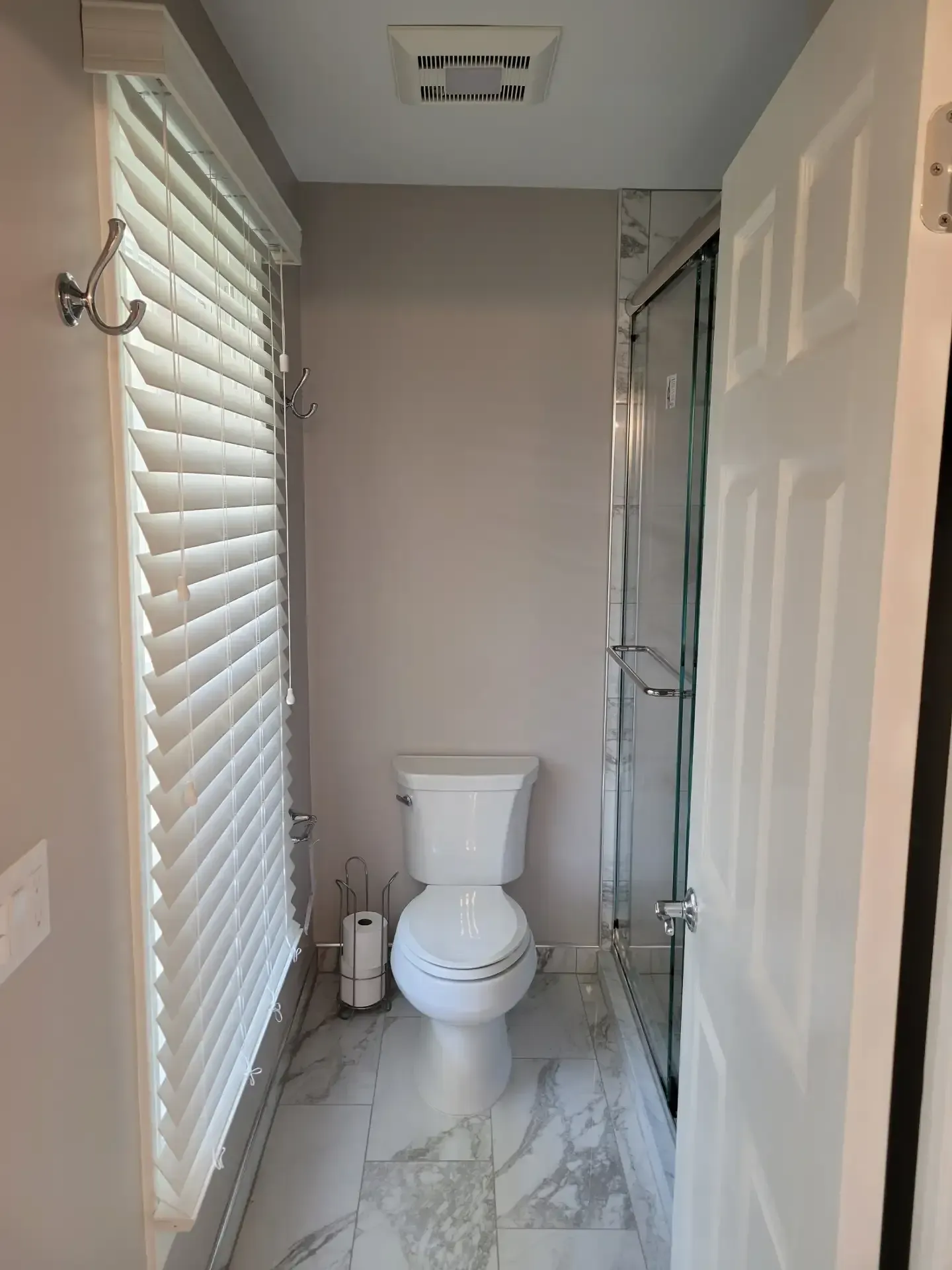 Small, neutral-toned bathroom with toilet, shower, blinds, and open door. Gray and white color scheme.