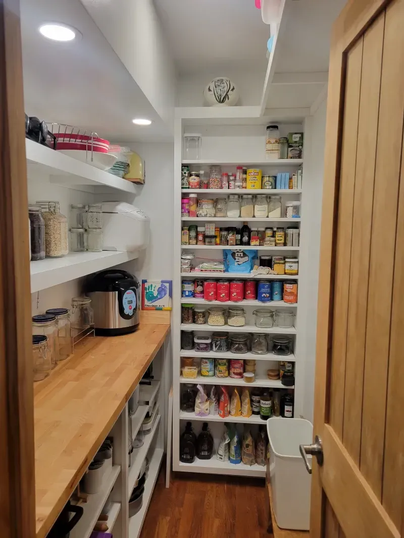 Pantry with white shelves and wooden countertop filled with food items. Door on right.