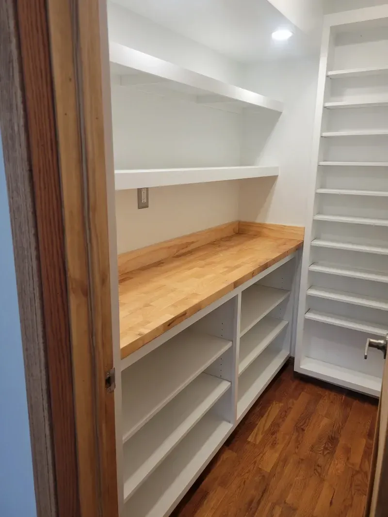 Pantry with white shelving, a wood countertop, and a tall shelf unit on the right. Wooden floor.