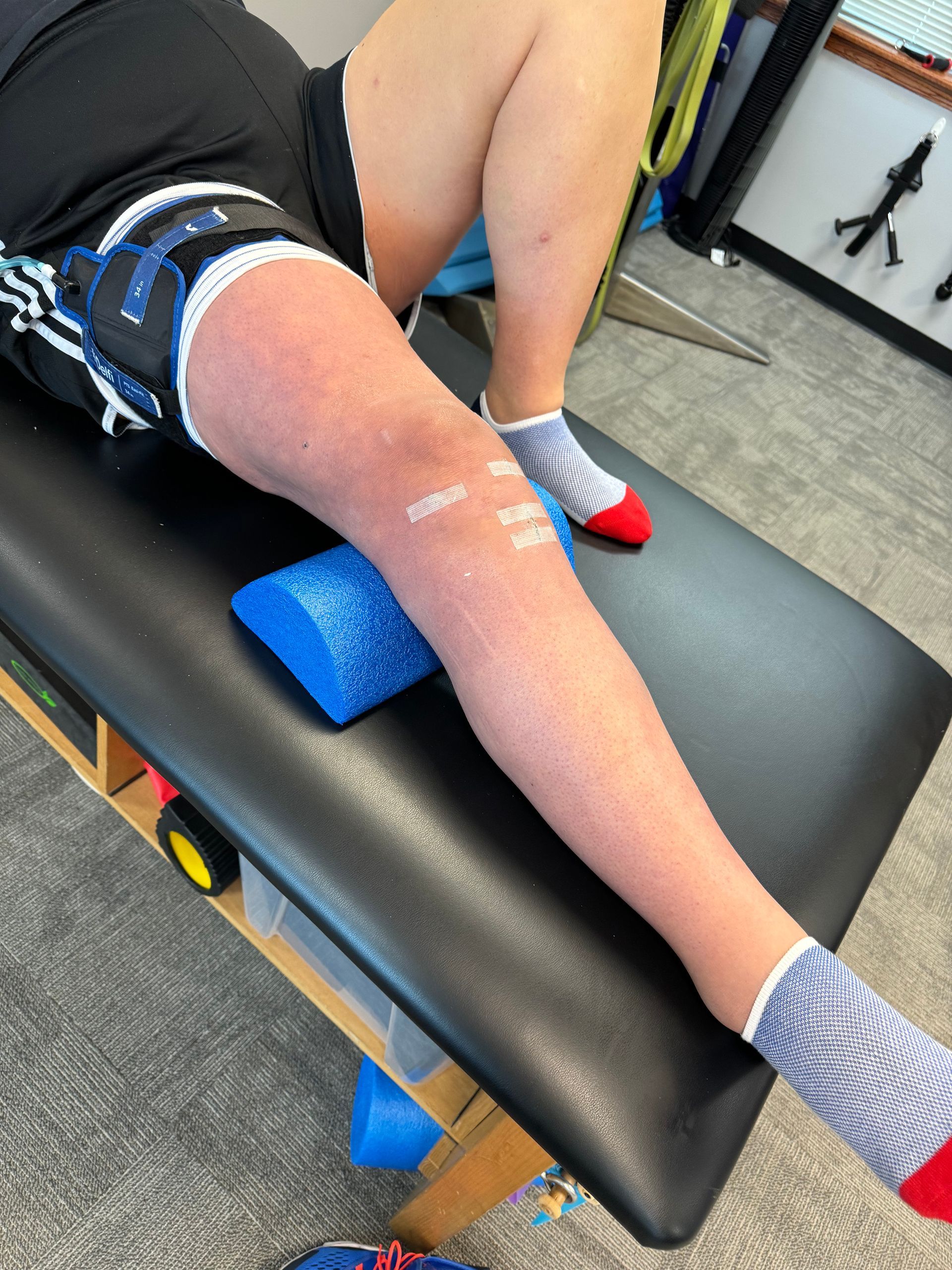 Person stretching on a therapy table with a blue foam pad under one knee and tape on the leg.