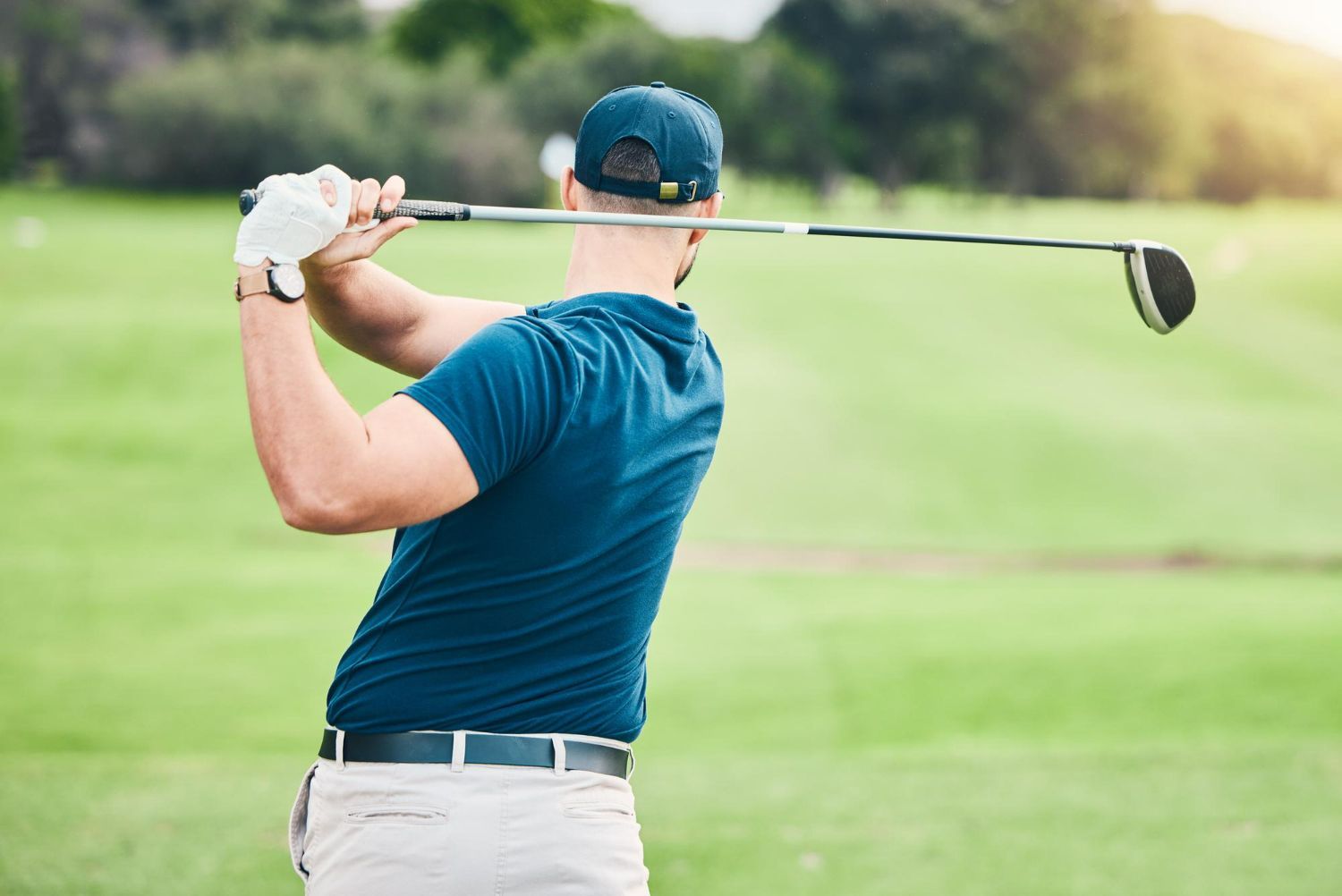 A golfer in a blue shirt and cap completes a swing on a green course.