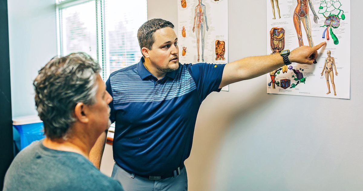 Two men in a clinic, one pointing at a medical chart on the wall.