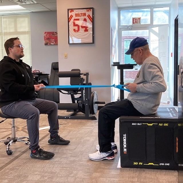 Two men seated indoors pulling a blue resistance band in a rehab or exercise room