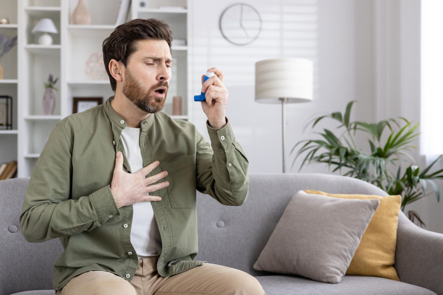 Man holding inhaler and clutching chest on couch in a bright living room