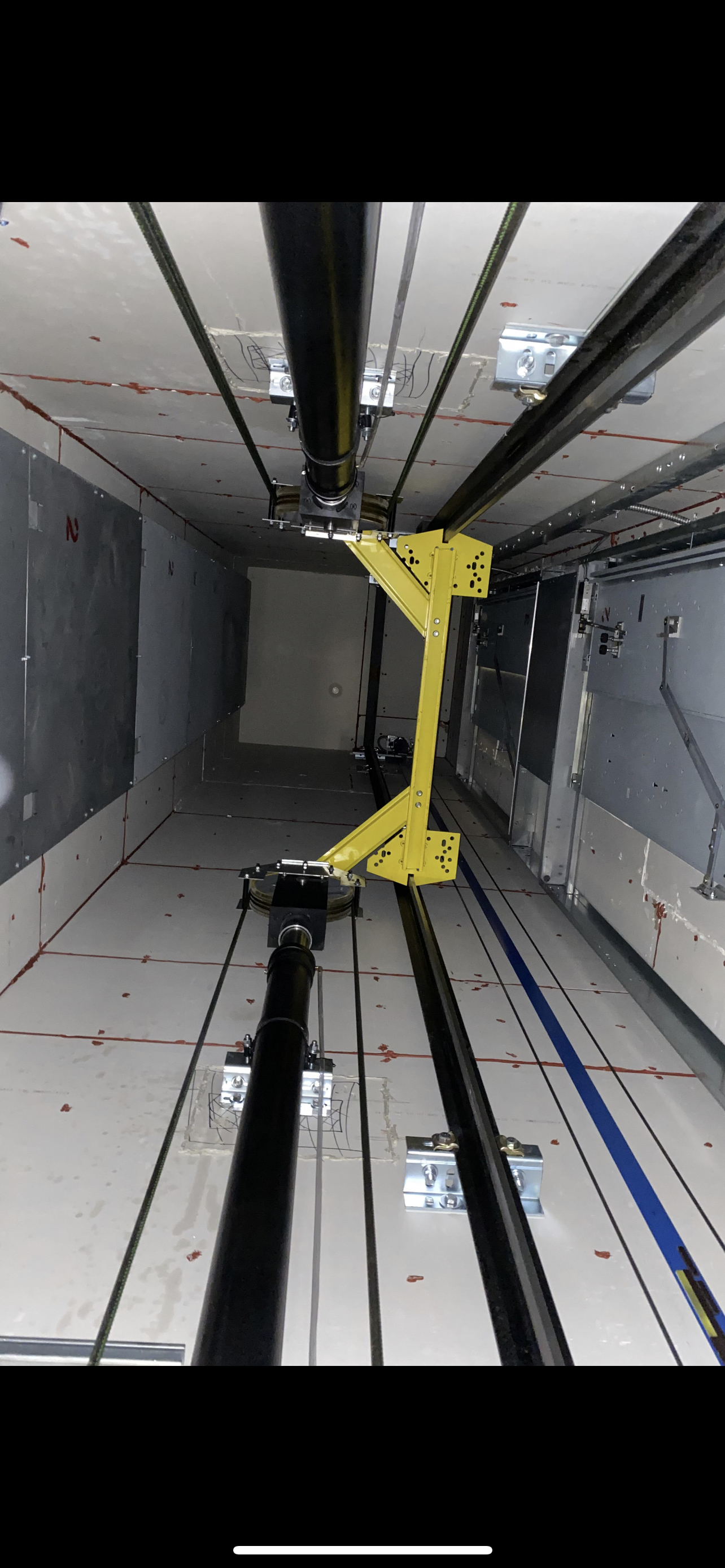 Intérieur de la cage d'ascenseur avec poutre de sécurité jaune. Câbles et rails visibles.
