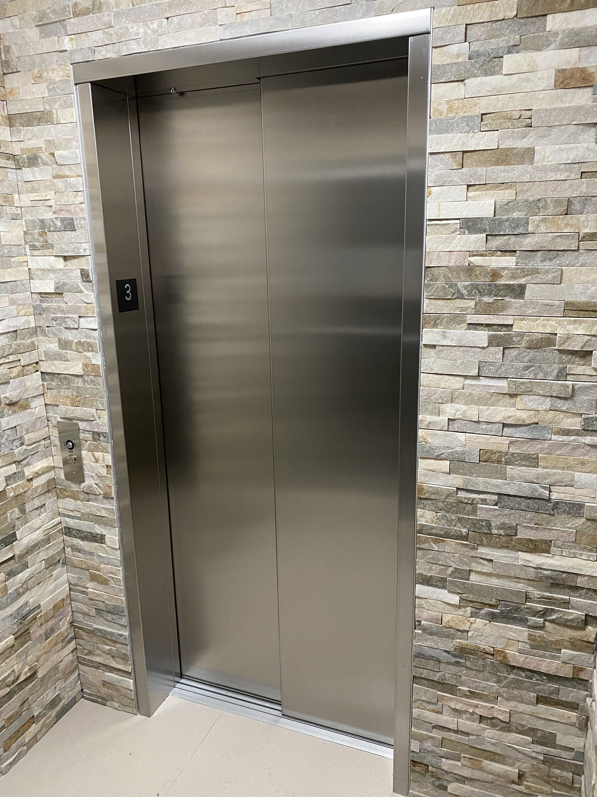 Ascenseur avec portes fermées en acier inoxydable encastrées dans un mur en pierre, boutons sur le côté gauche, un 3 est affiché.
