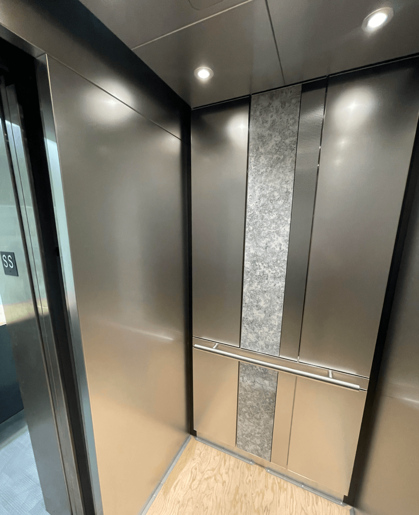 Intérieur d'ascenseur vide avec parois en acier inoxydable, rampes et moquette. Flèche rouge vers le haut allumée.