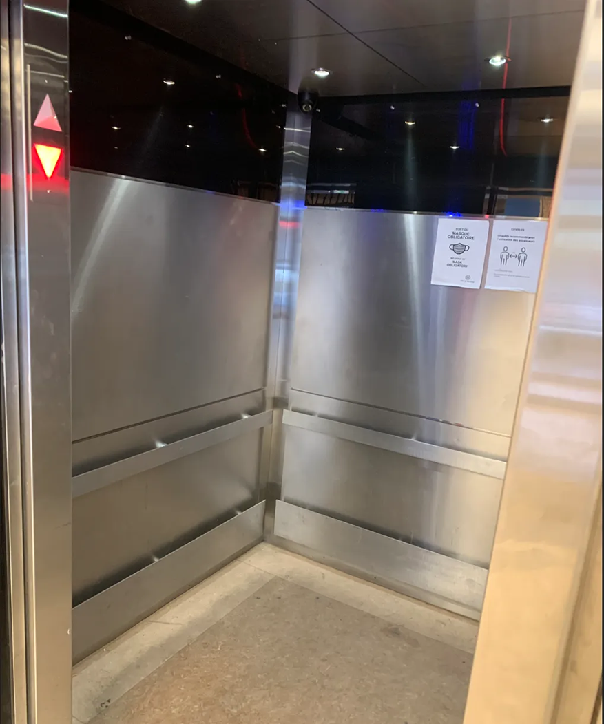Vue intérieure d'un ascenseur avec des parois en acier inoxydable et un sol gris.